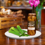 A jar of Hot Pickled Okra on white plate with okra and green chili pepper