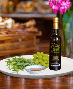 A 200ml bottle of Tarragon White Balsamic Vinegar on white plate with tarragon leaves and sample balsamic vinegar